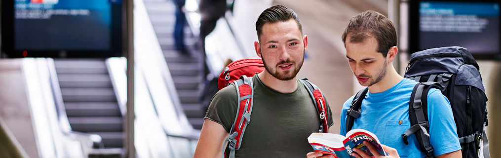 Two male travelling companions looking through a travel guide at the bottom of an escalator.