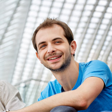 A young man sitting inside a glass ceilinged building smiling.