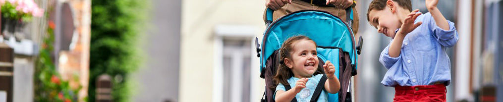 Young siblings walking happily down the street, one being pushed in a stroller and the other walking along side her. 