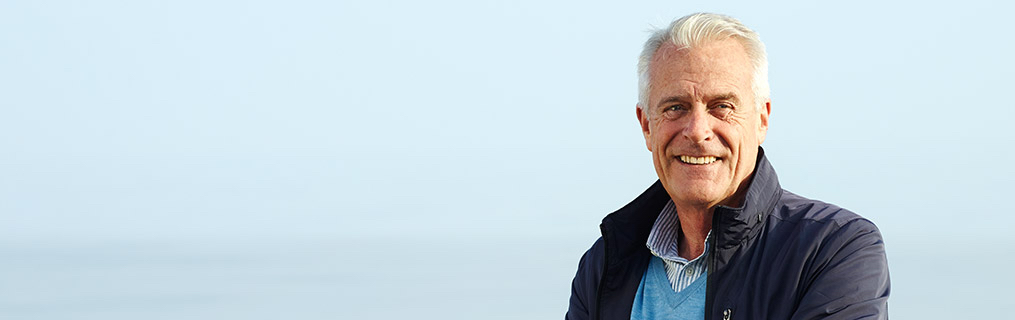 A man in his early sixties standing and smiling in front of a clear, blue sky.