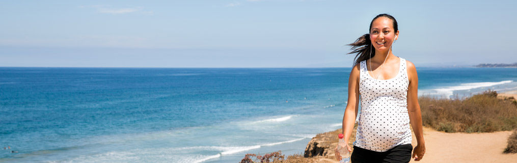 A pregnant woman happily walking down a sunny beach.