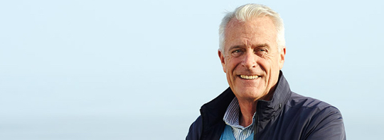 A man in his early sixties standing and smiling in front of a clear, blue sky. A man in his early sixties standing and smiling in front of a clear, blue sky.